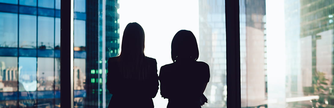 Two professional women looking outside a large window.